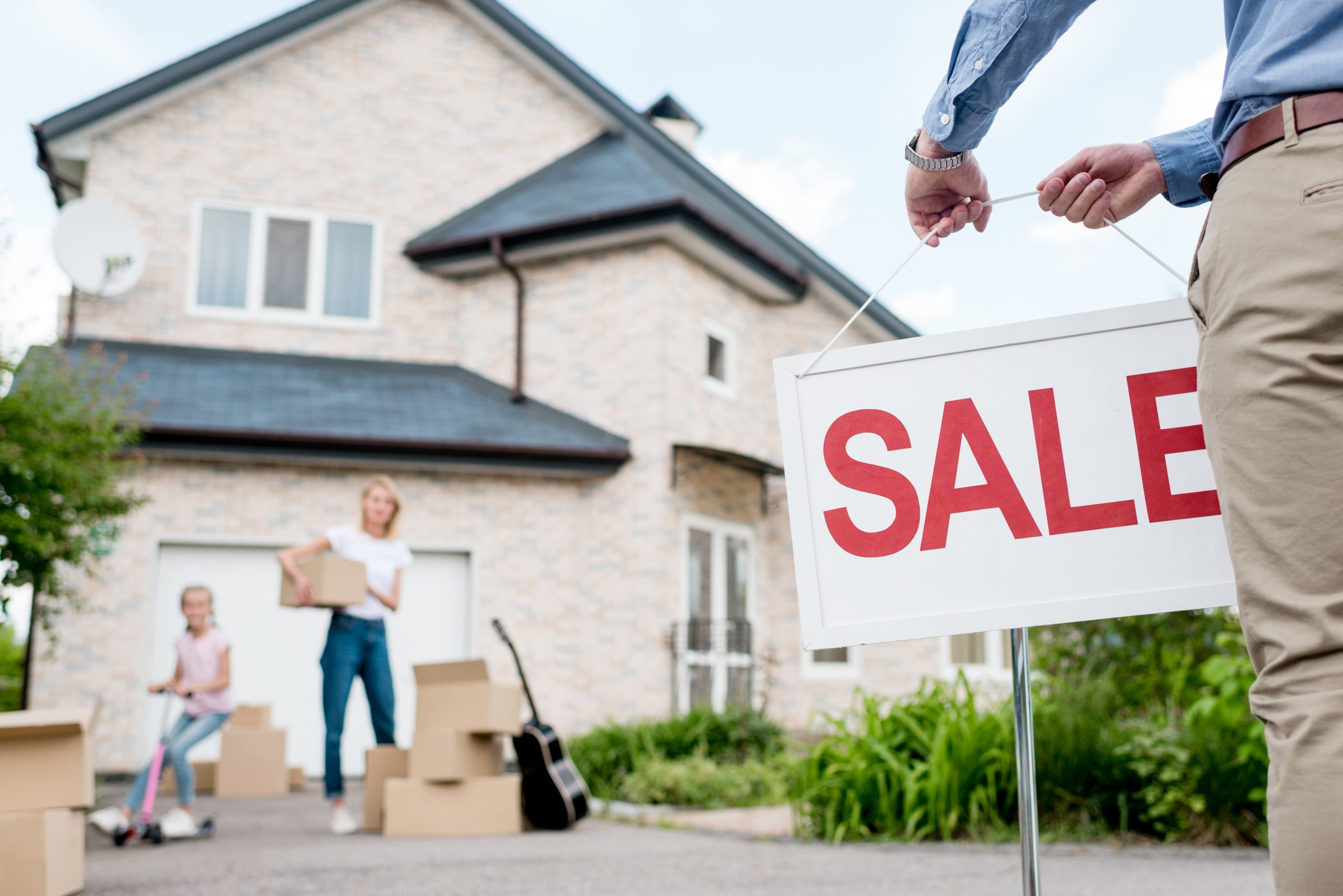 Cropped image of male realtor with sale signboard and family moving in with cardboard boxes behind