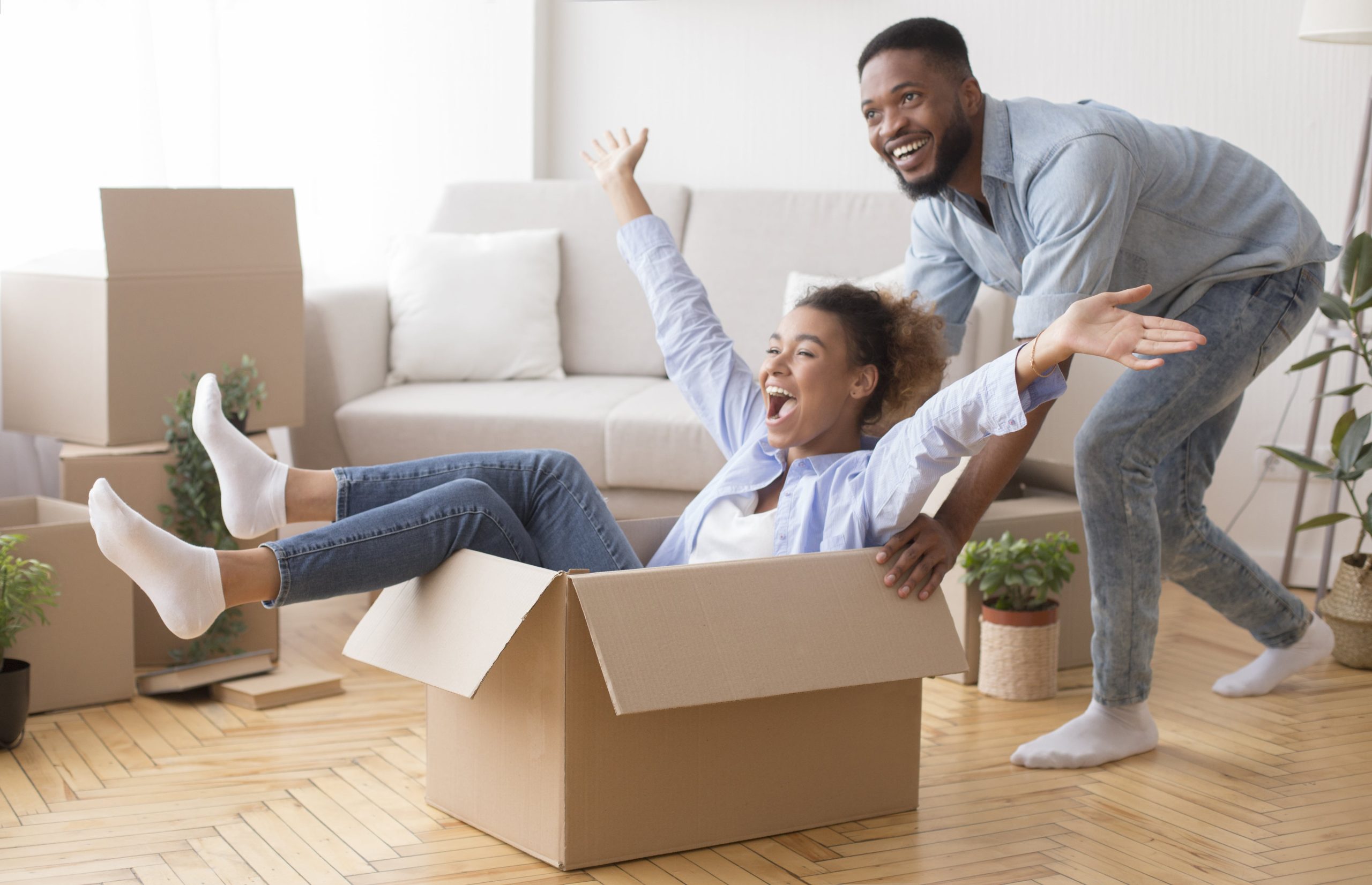 Excited African American Man Pushing Woman Riding In Cardboard Box Celebrating Moving Into New House
